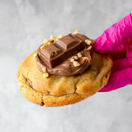 Wholenut Deluxe Cookie with chocolate frosting and KitKat bar held by a pink-gloved hand on a light gray background