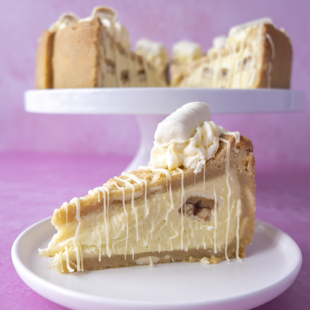 A slice of Cookie Pie with White Twix with white chocolate drizzle sits on a small white plate in the foreground, with a larger full pie on a white serving stand blurred in the background against a pink-purple backdrop.