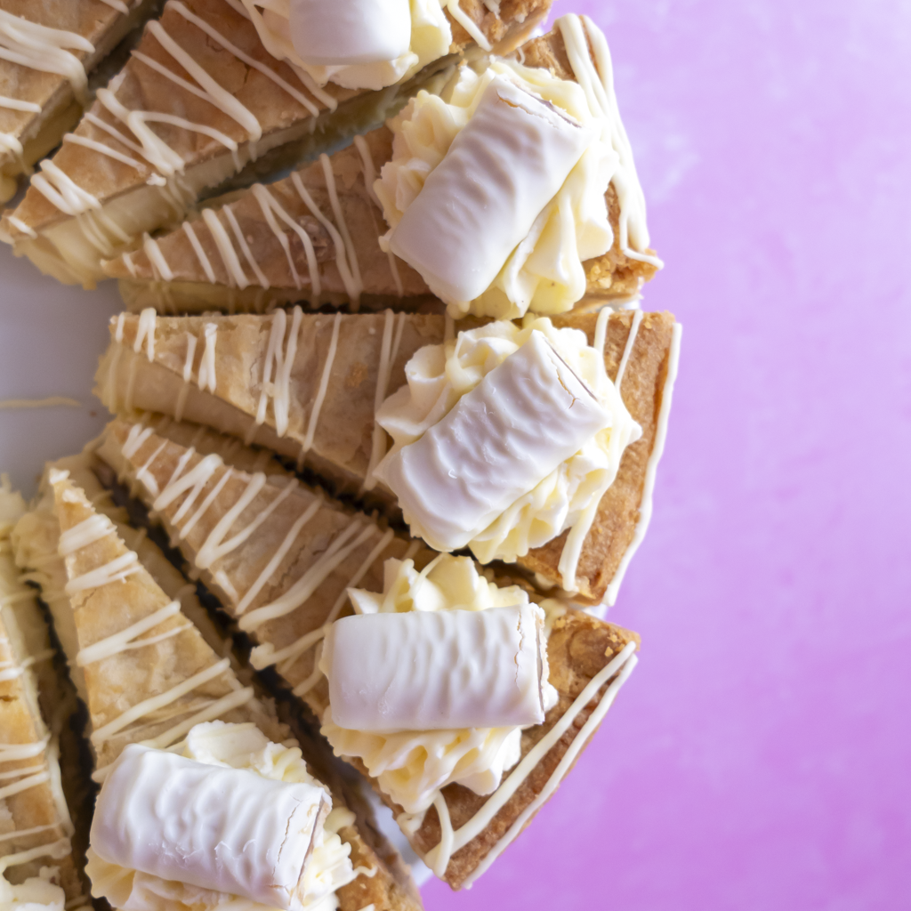 Slices of a Cookie Pie with White Twix topped with white cream and drizzled with white chocolate, arranged on a white surface against a light pink background.