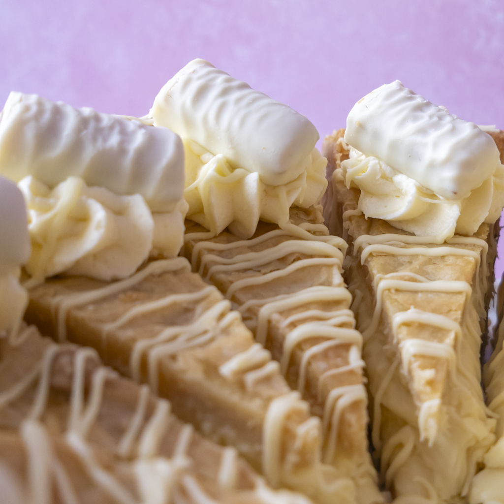 Close-up of several triangular slices of Cookie Pie with White Twix, topped with piped white cream and drizzled with white chocolate, set against a soft pink background.