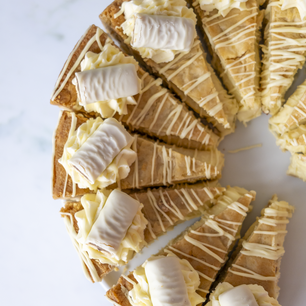 A top-down view of a Cookie Pie with White Twix cut into triangular wedges, topped with white cream and drizzled with white chocolate on a light marble surface.
