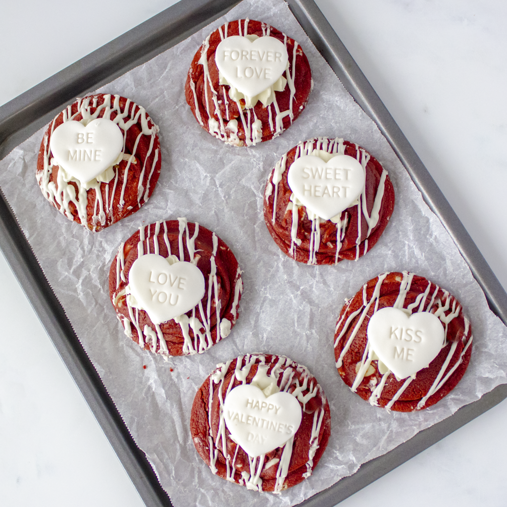 6 Red velvet cookies with white heart-shaped messages on a gray baking tray.