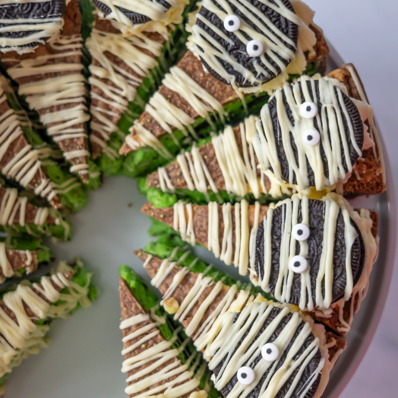 A Halloween-themed cookie pie cut into slices, decorated with Oreo cookies, white chocolate drizzle, and small candy eyes, served on a light grey plate.