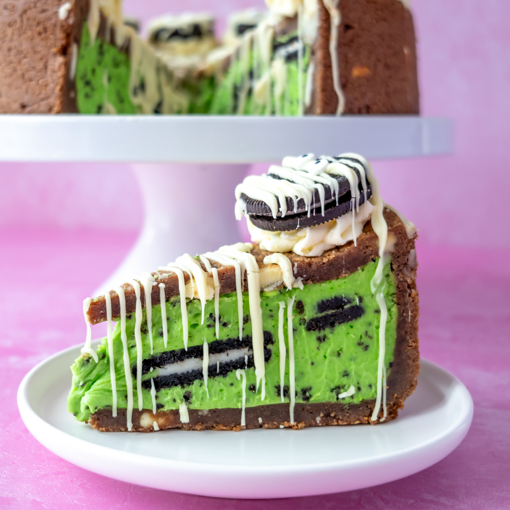 A slice of green Midnight Monster Cookies n Cream Pie with Oreo cookie pieces and white chocolate drizzle, served on a small white plate with a blurred whole cake on a stand in the background against a pink backdrop.