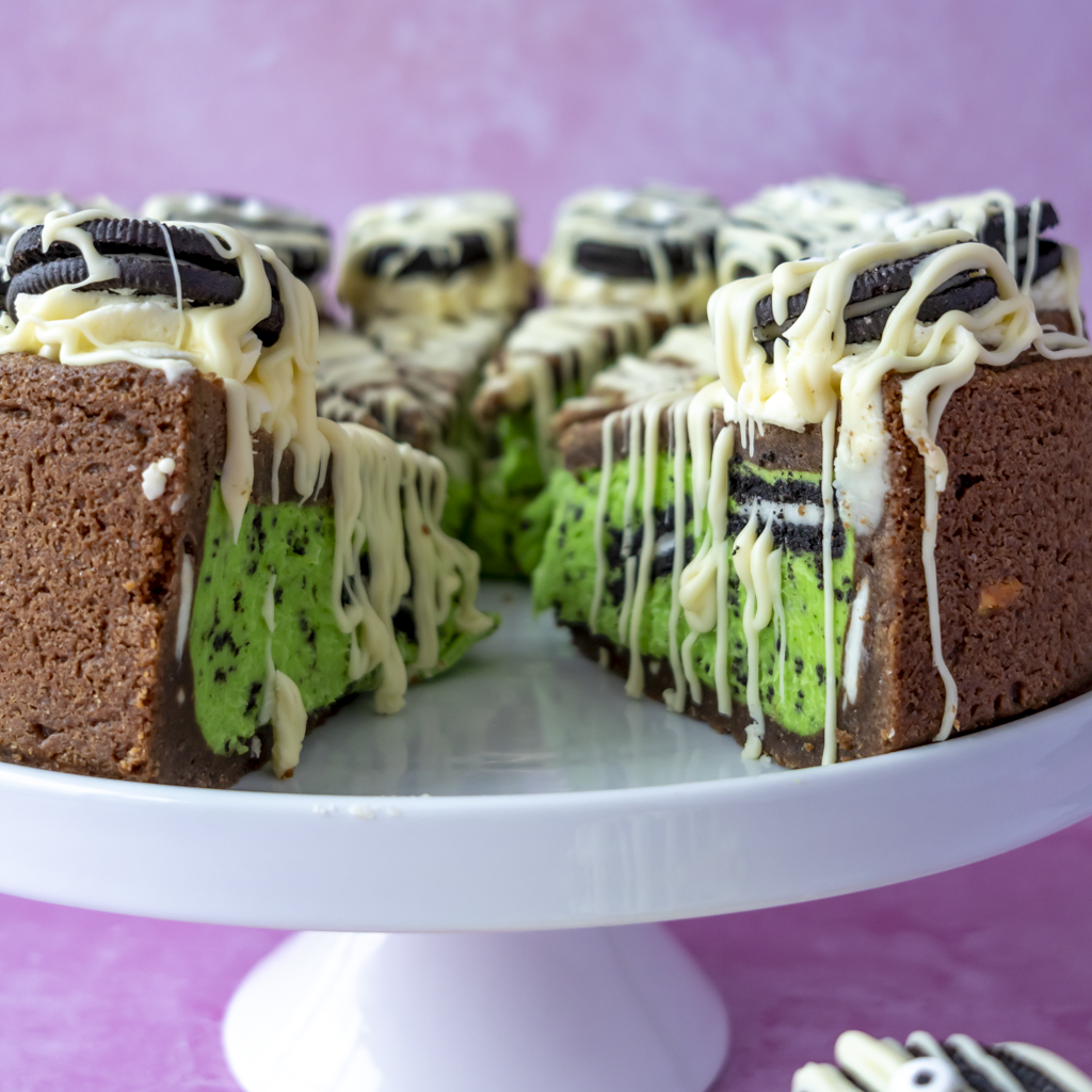Chocolate cake slices with bright green filling and white chocolate drizzle, topped with Oreo cookies, displayed on a white cake stand against a pink background.
