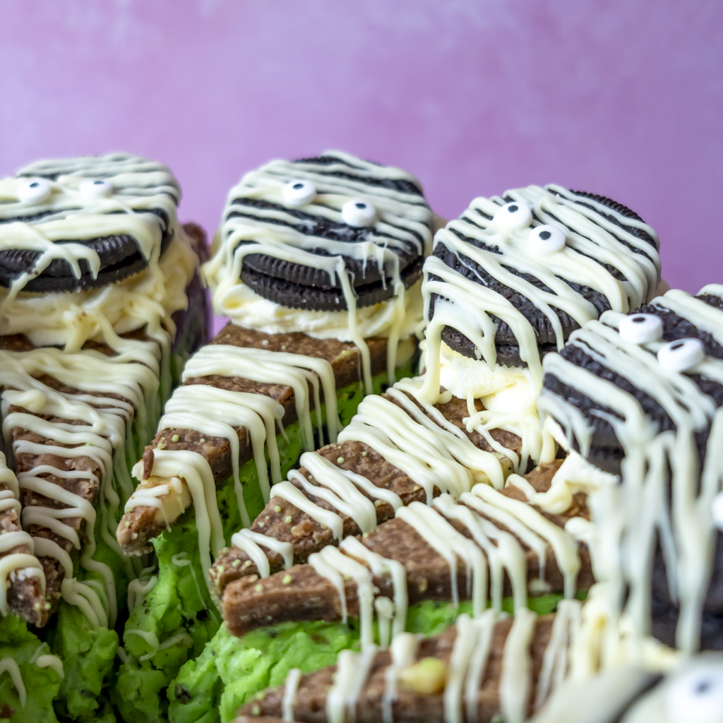 Mummy-themed cookie pie slices decorated with white chocolate drizzle, green chocolate filling, and sweet eyes, arranged on green frosting with Oreo cookies in the background against a pink backdrop.