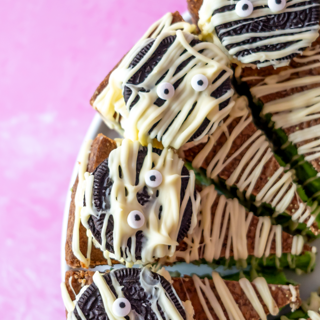 Mummy-themed Oreo cookies decorated with white chocolate drizzle and sweet eyes, arranged on a plate against a pink background.