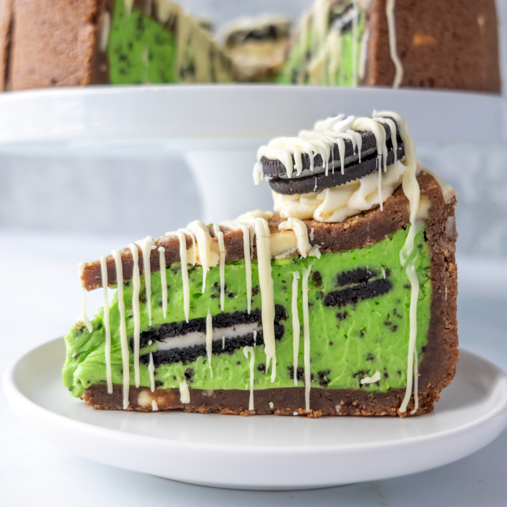A slice of green cookie pie dessert on a white plate, featuring a chocolate cookie crust, bright green pie filling with cookie crumbles, white chocolate drizzle, and a cookie topping on a cake stand.
