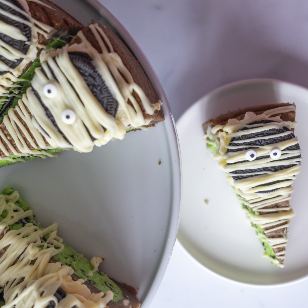 A Halloween-themed slice cookie pie decorated with Oreo cookie faces and white drizzle, shown on a round cake pan with a single slice served on a small plate.