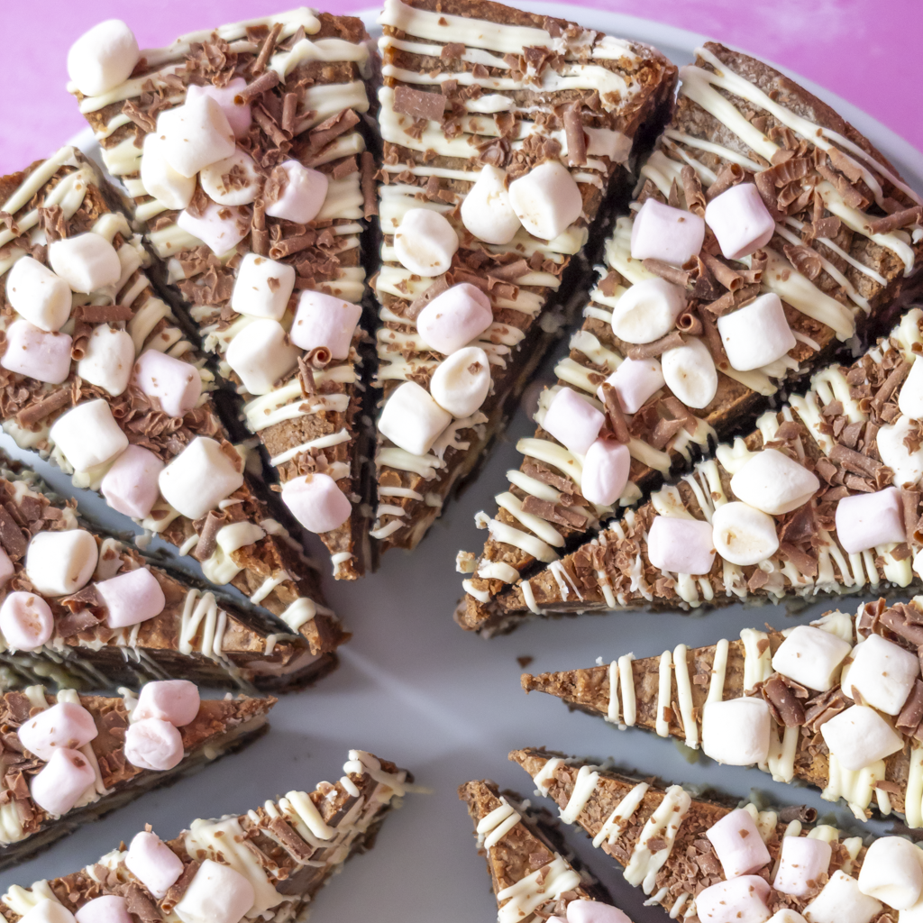Chocolate and white chocolate cookie pie slices topped with mini marshmallows and chocolate shavings, arranged on a white plate against a pink background.