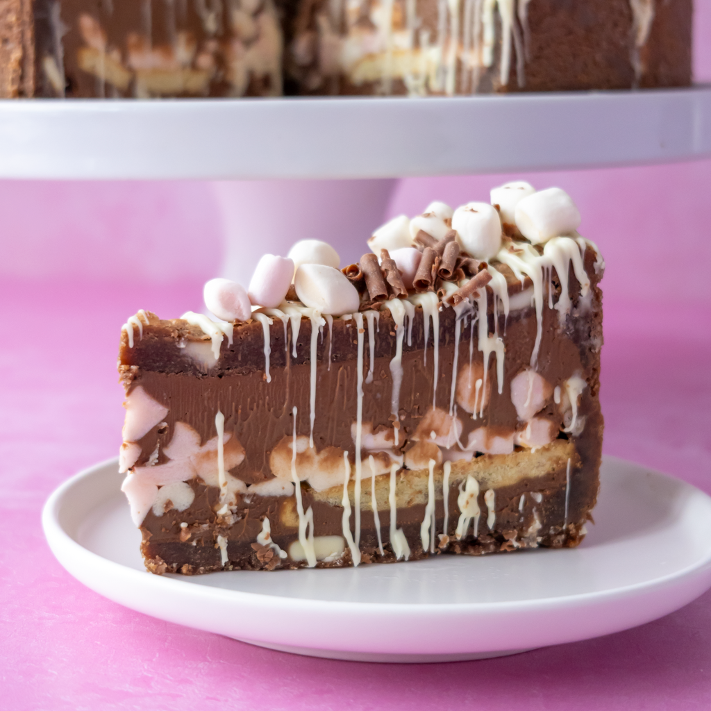A slice of chocolate cookie pie on a white plate against a pink background, topped with white chocolate drizzle, mini marshmallows, and chocolate curls.