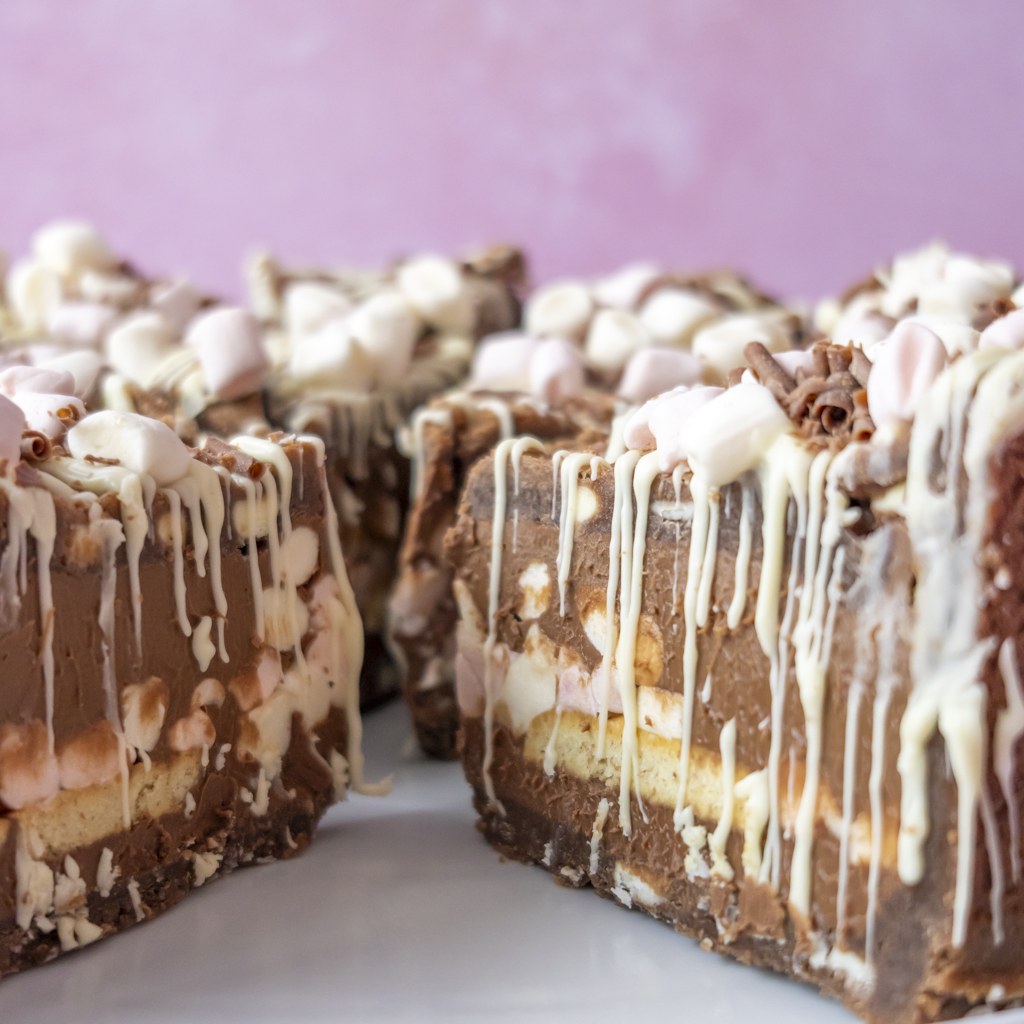 S’mores Cookie Pie topped with white chocolate drizzle and mini marshmallows, arranged on a white plate against a soft pink background.