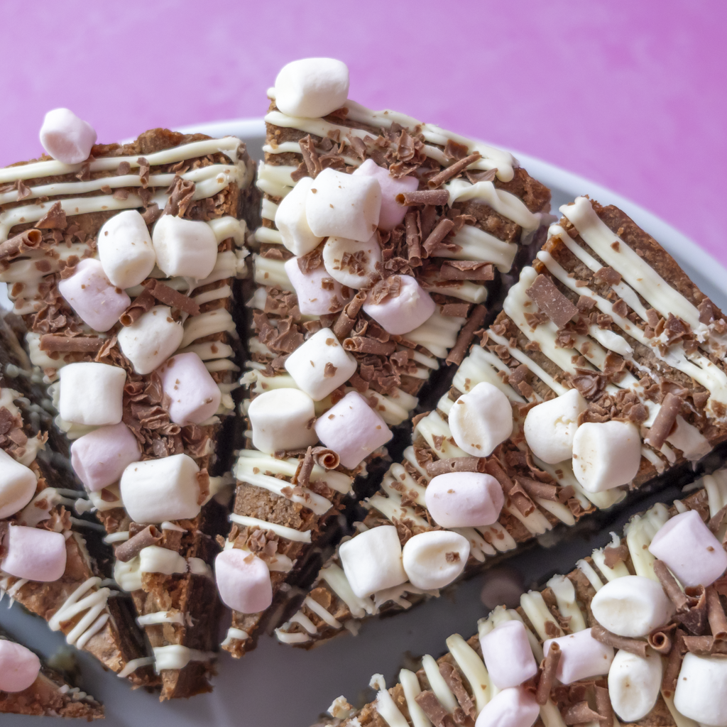 Slices of S’mores Cookie Pie topped with white chocolate drizzle, chocolate shavings, and pink and white marshmallows, arranged on a white plate against a pink background.