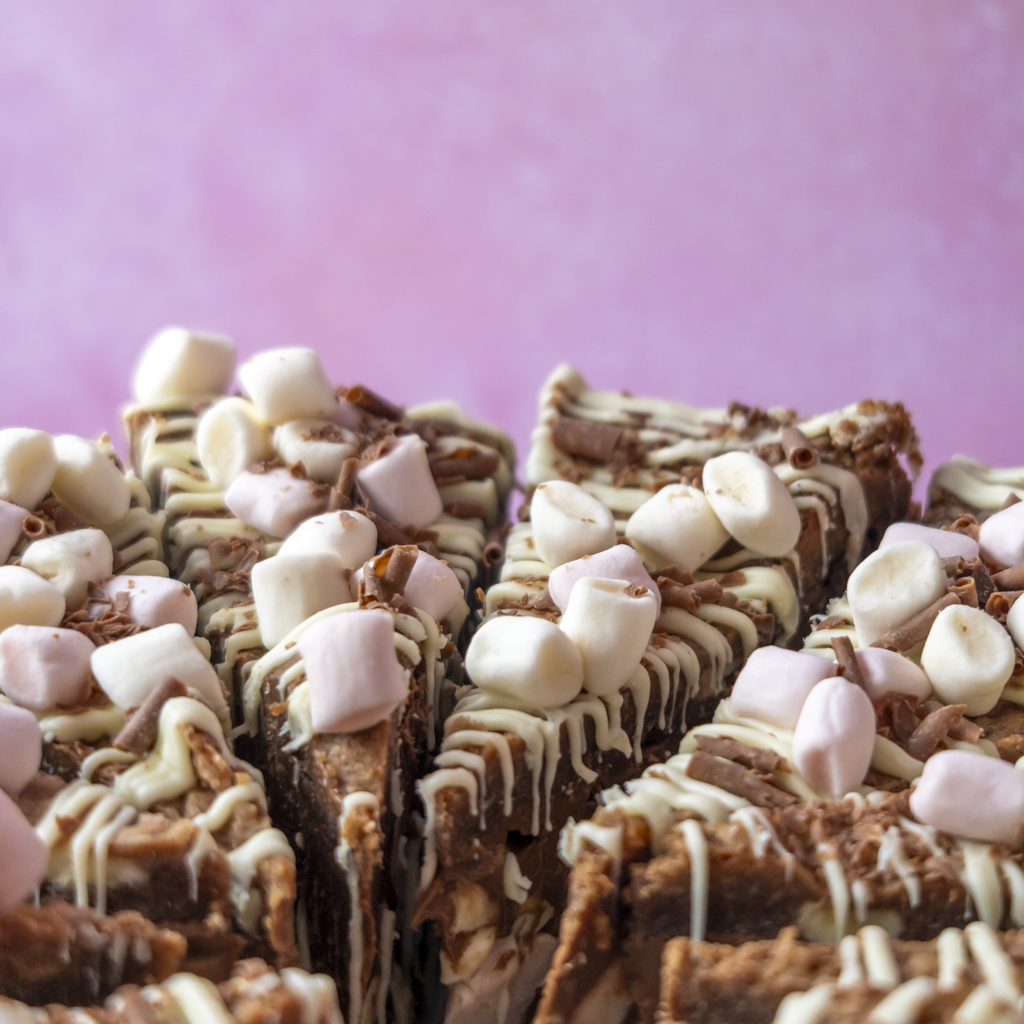 Close-up of S’mores Cookie Pie topped with white chocolate drizzle, mini marshmallows, and chopped nuts, arranged against a soft pink background.