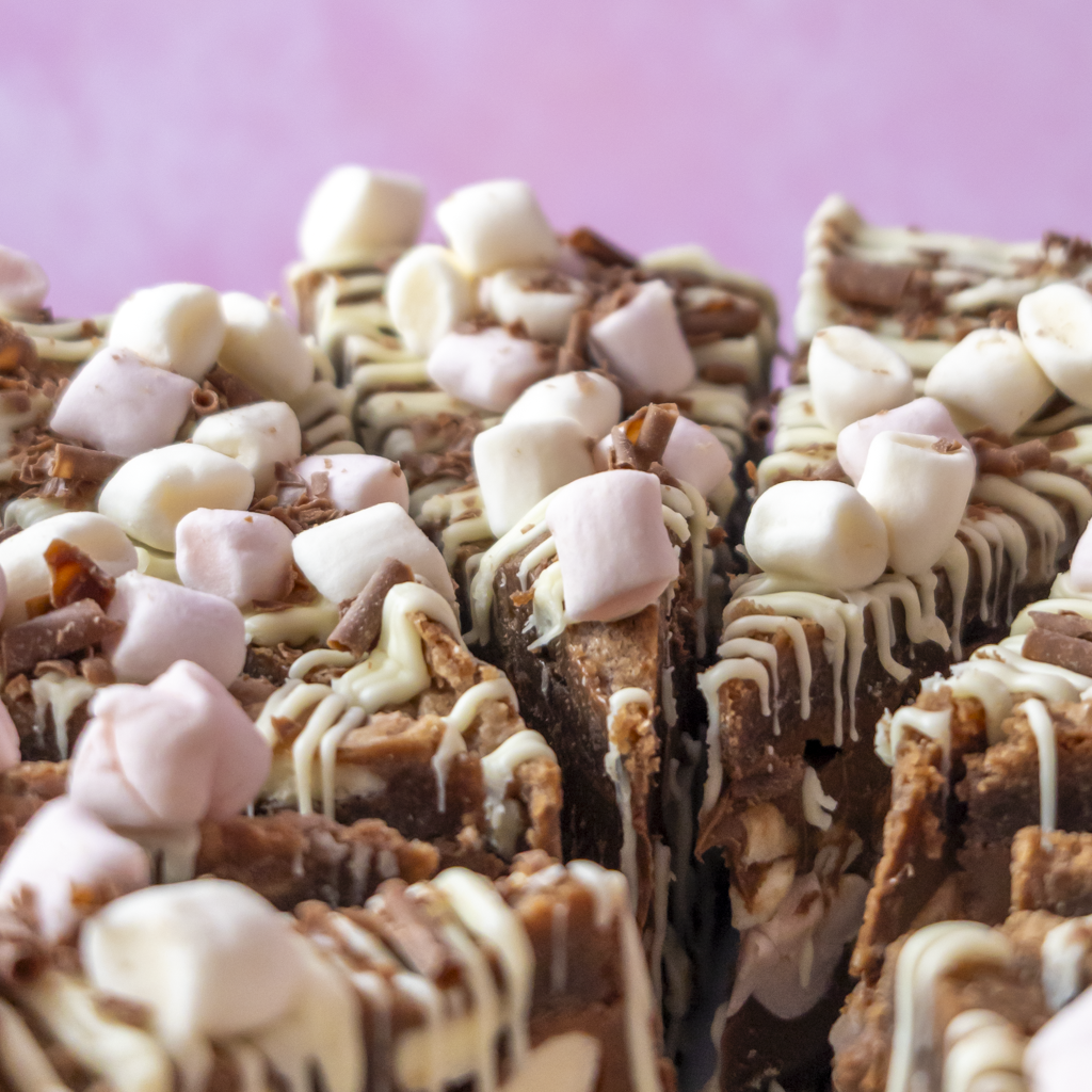 Close-up of chocolate-covered S’mores Cookie Pie topped with mini marshmallows and white chocolate drizzle, set against a soft pink background.